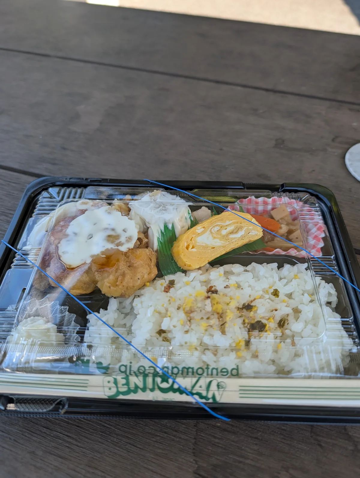 An open Bentman bento box on a wooden table with rice, shumai, tamagoyaki, fried fish, and pickled vegetables, secured with a blue rubber band