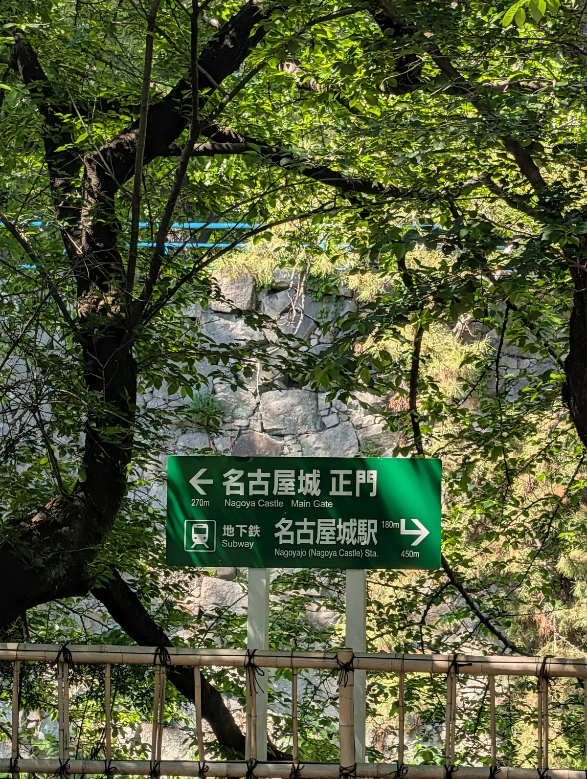 Bilingual Japanese-English wayfinding sign for the Nagoya Castle main gate and Nagoyajo subway station, framed by lush green trees
