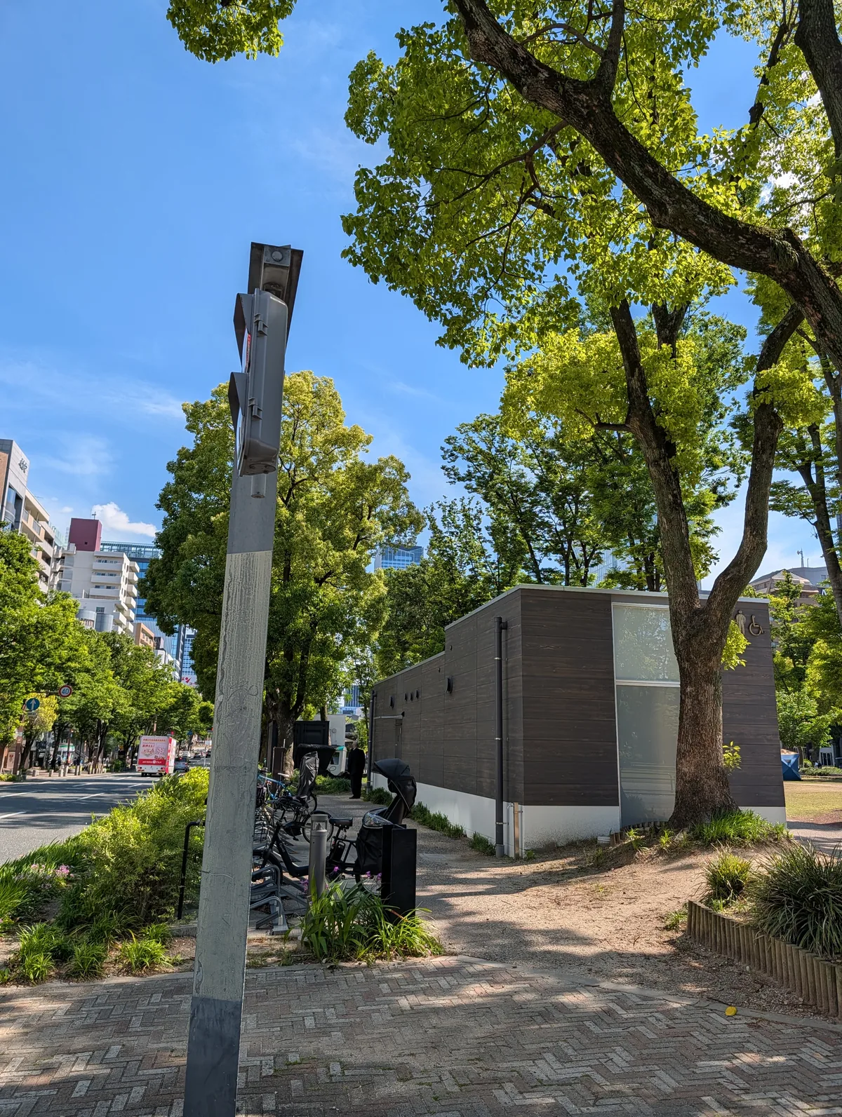 A bicycle parked beside a small public restroom building under tall green trees, on a brick-paved path through a city park near Nagoya Castle