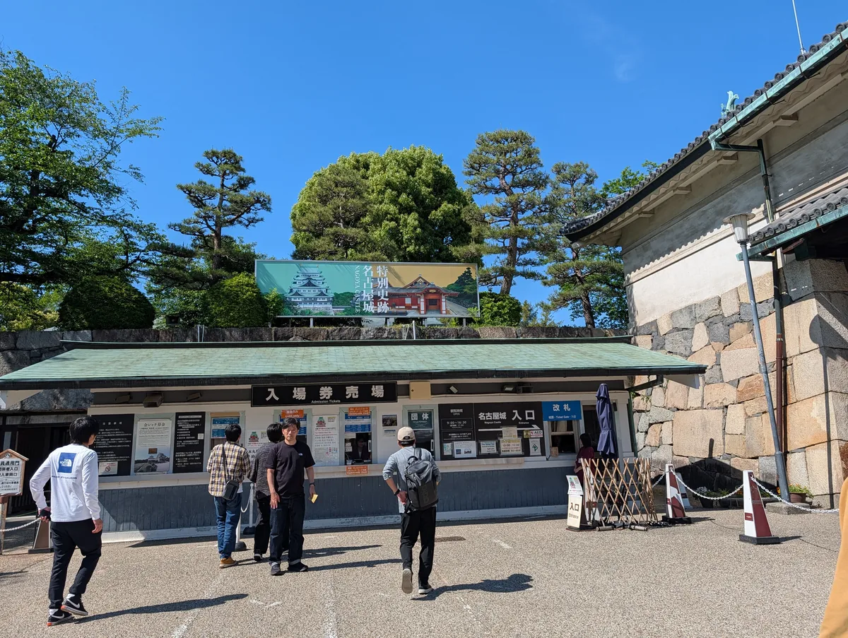 The Nagoya Castle ticket booth on a sunny May day, with a small queue of visitors and pine trees and the castle skyline in the background