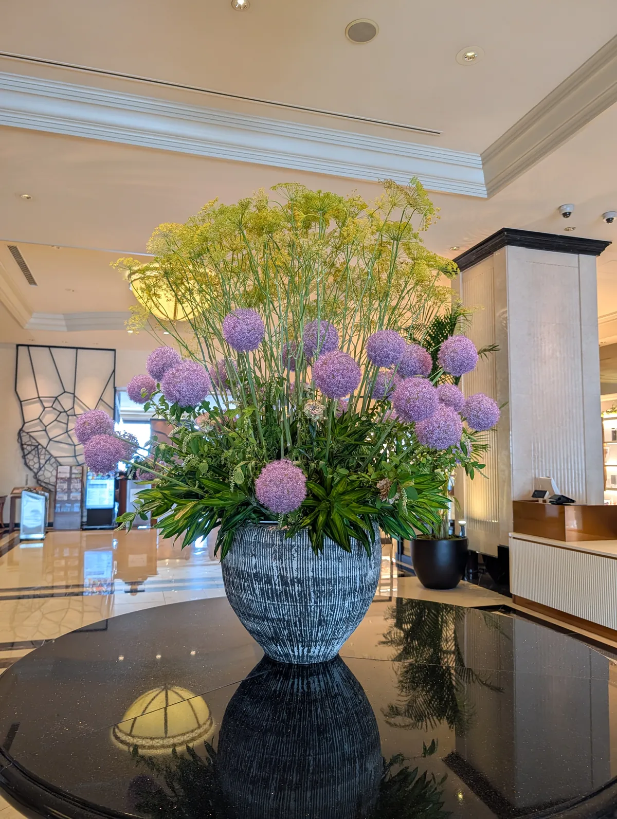 A large purple allium and dill flower arrangement in a tall blue-grey vase on a polished black stone pedestal in the Marriott Associa lobby