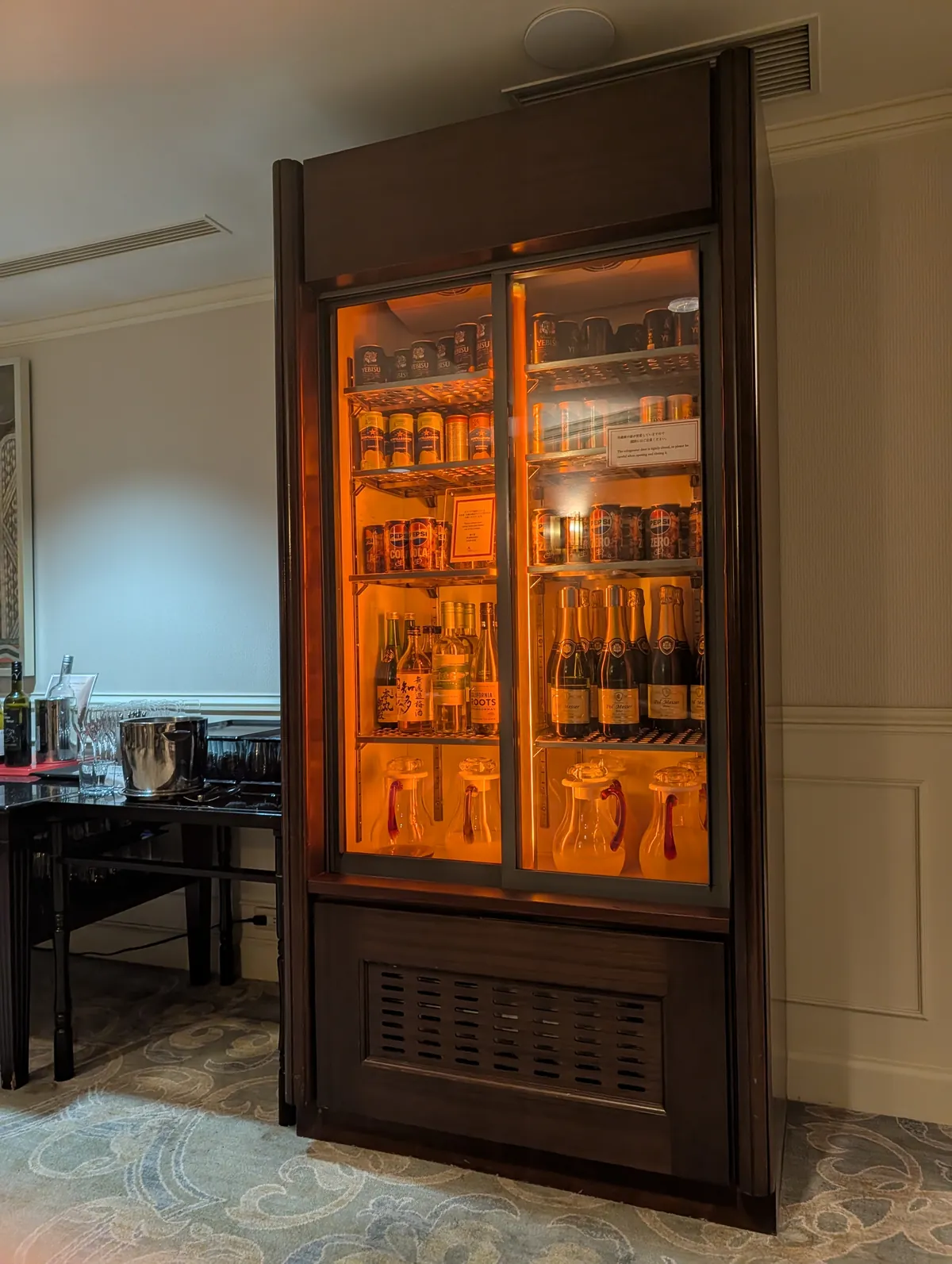 Lit display cabinet inside the Marriott Associa executive lounge stocked with champagne bottles, sake, and Japanese craft beer cans