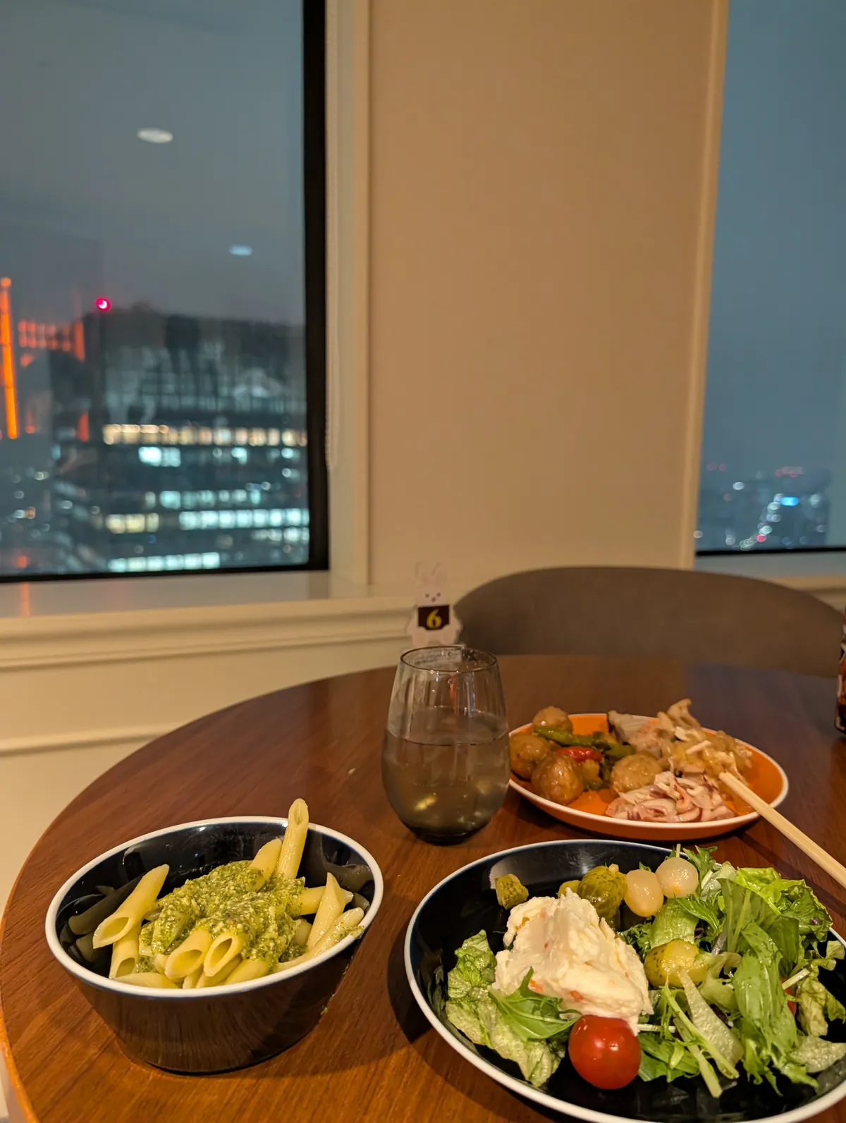 An evening table inside the Marriott Associa executive lounge with a bowl of pesto penne pasta and a salad, with the lit Nagoya skyline visible through the high-floor window