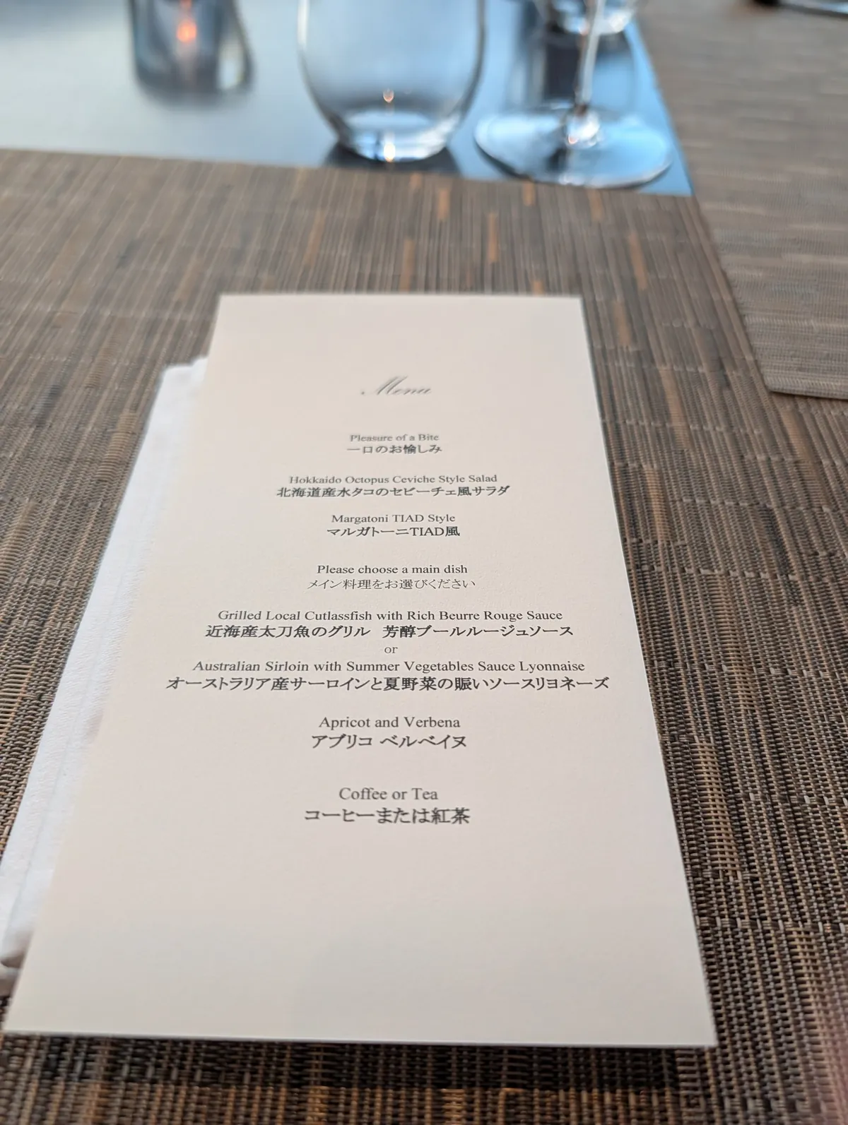 The bilingual TIAD dinner menu card on a textured placemat showing courses including Margatoni TIAD Style and a choice of grilled cutlassfish or Australian sirloin