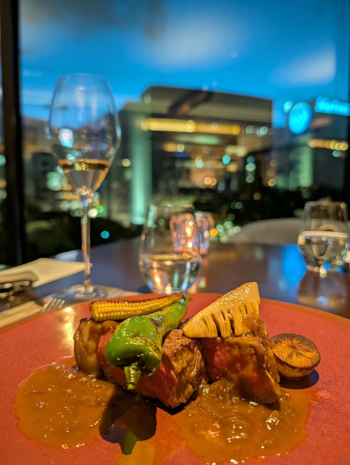 A grilled Australian sirloin main course on a red plate with charred shishito pepper, baby corn, and bamboo shoot, with the Nagoya skyline lit up at dusk through the restaurant window