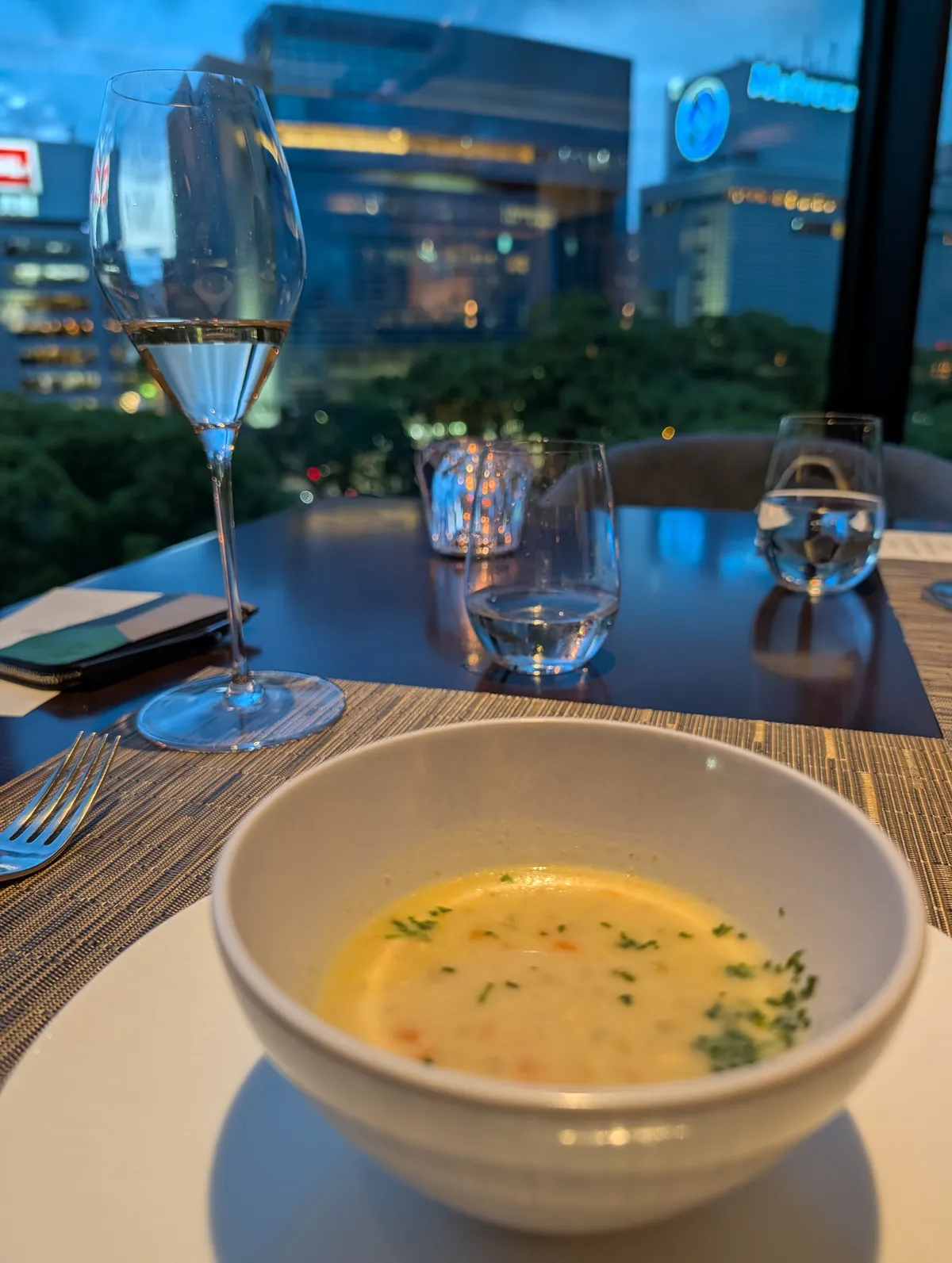 A bowl of cream soup with parsley alongside a glass of champagne at TIAD restaurant, with the Nagoya skyline visible at dusk through floor-to-ceiling windows