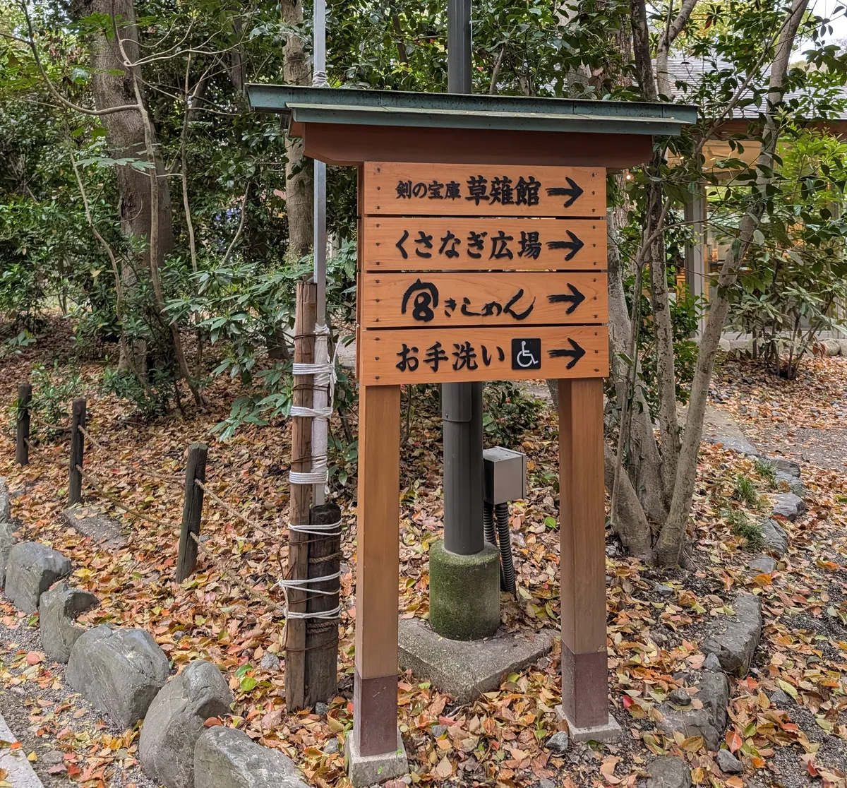 Wooden shrine signboard with four slats reading Kusanagi-kan sword museum, Kusanagi Plaza, Miya Kishimen and restrooms, on a path covered in fallen leaves