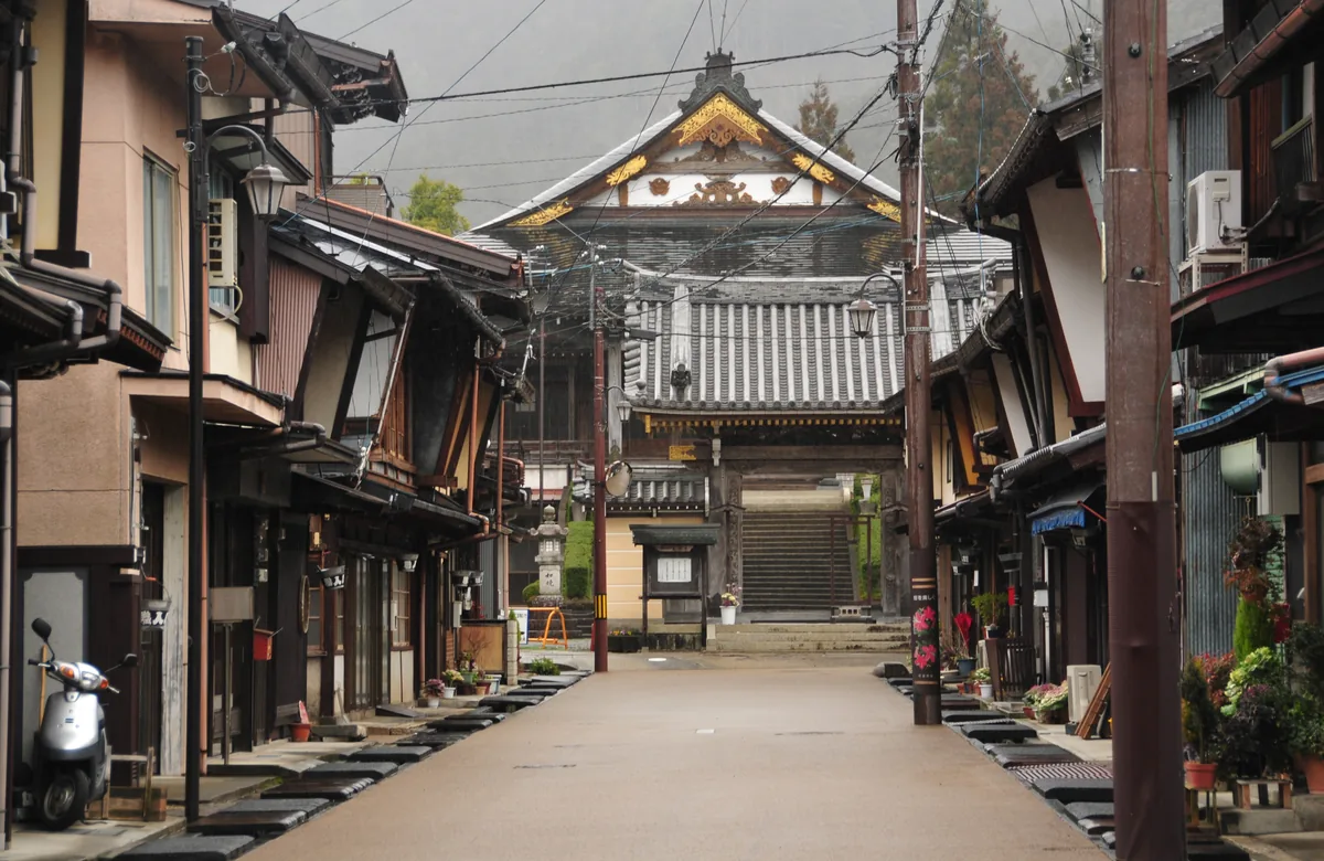 A quiet street of wooden townhouses leading to a temple gate at the end, with no people in sight
