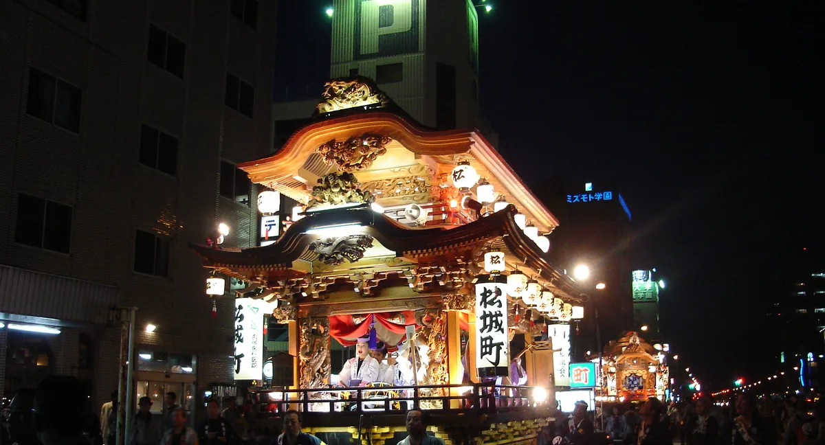 An illuminated gilded festival float hung with lanterns being pulled along a city street at night