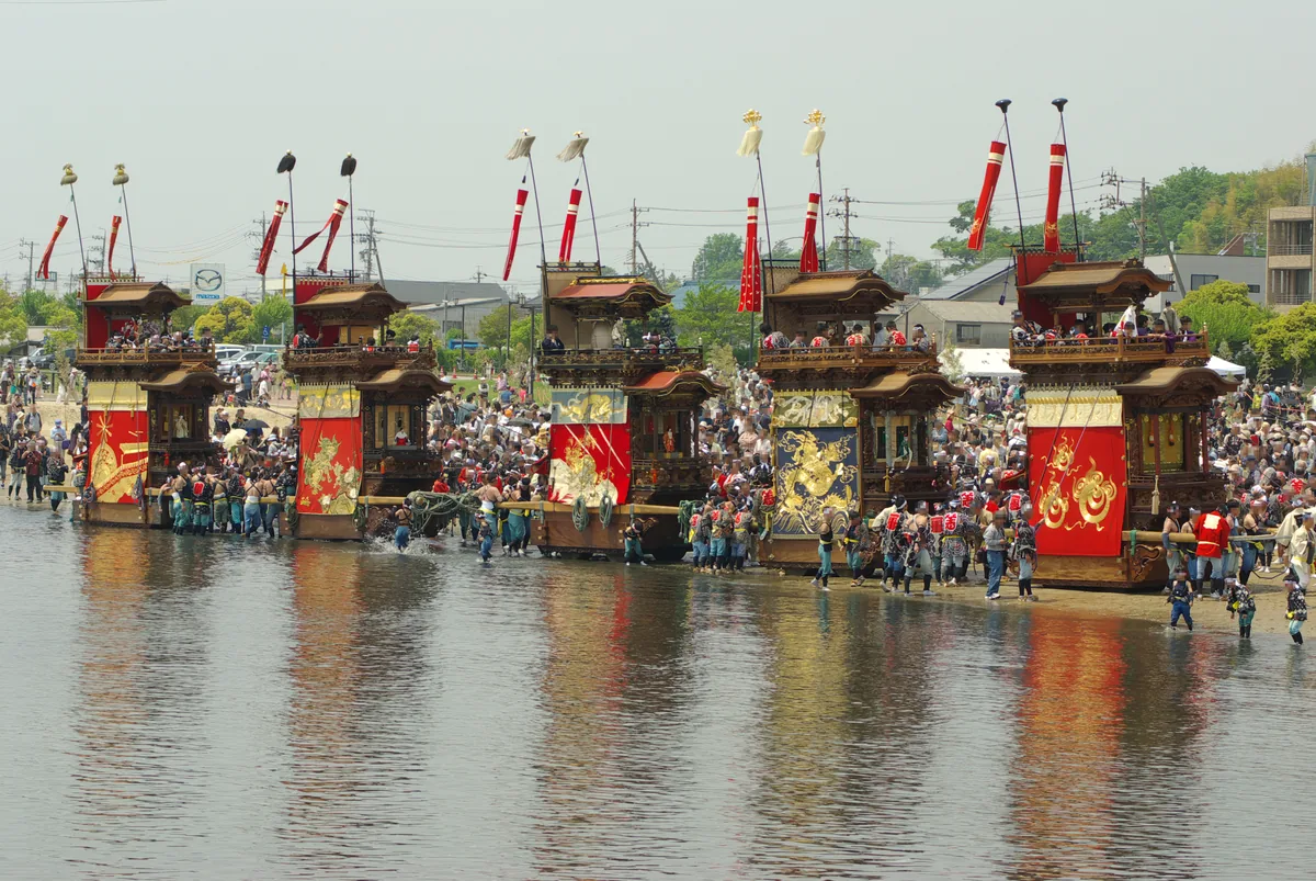 Five ornate wooden festival floats pulled through shallow tidal water by teams in happi coats