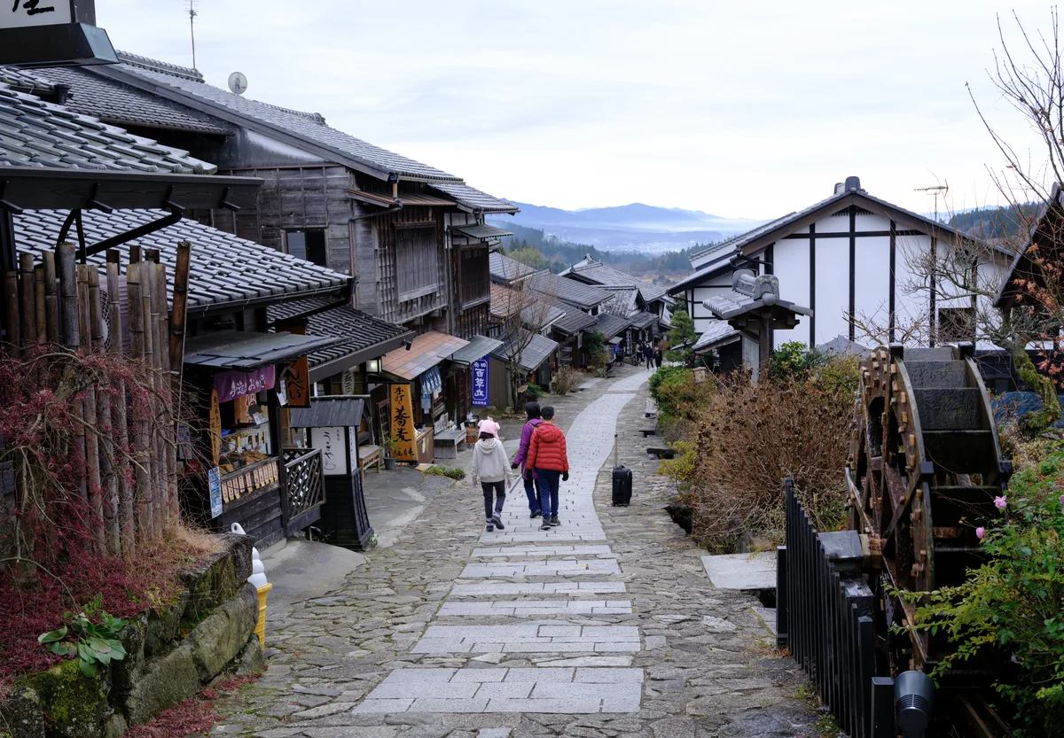 A stone-paved slope lined with old wooden post-town buildings and a water wheel, mountains beyond