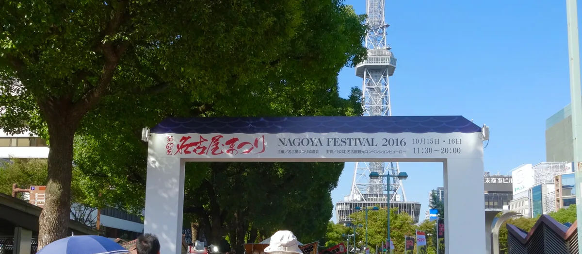 Festival gate with a banner reading Nagoya Festival, with the Nagoya TV Tower rising behind trees