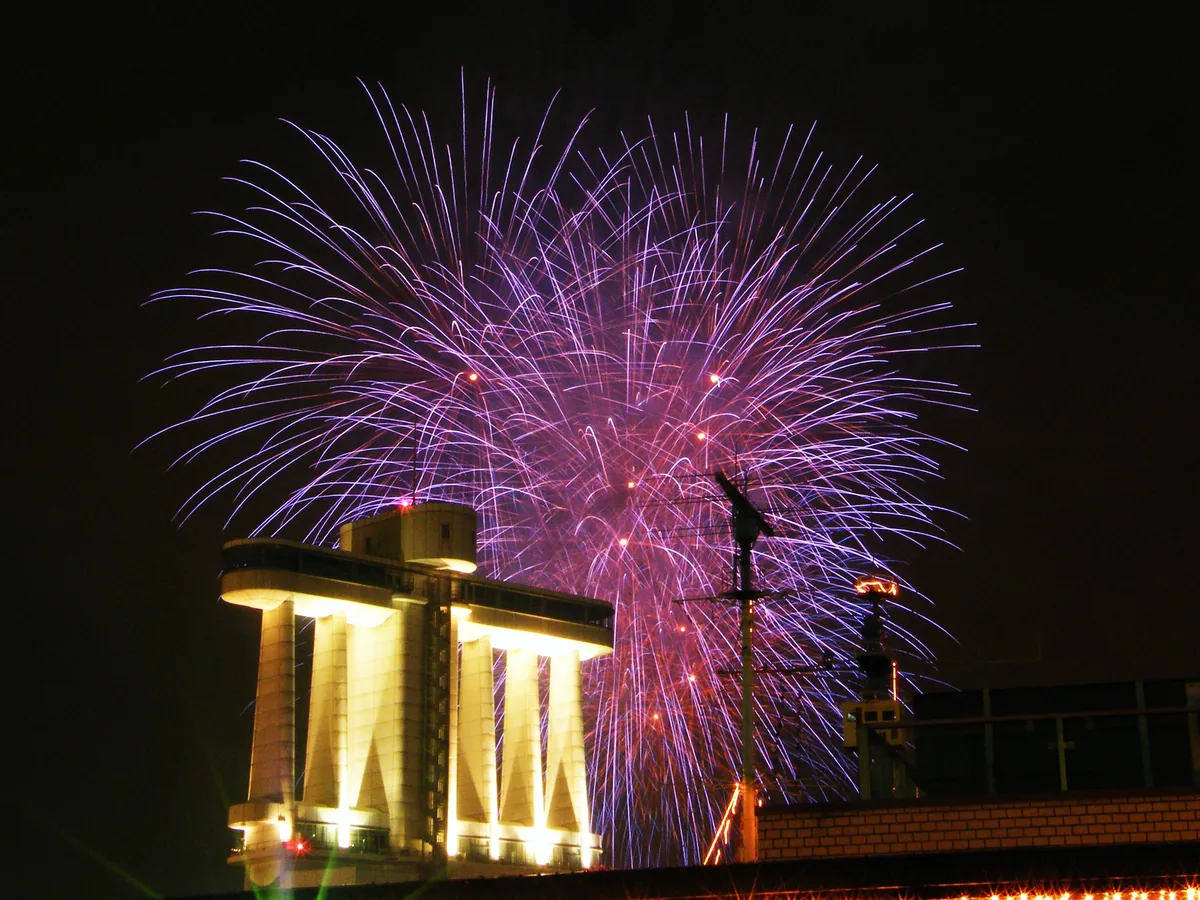 Purple fireworks bursting in the night sky above a floodlit pier building at Nagoya Port