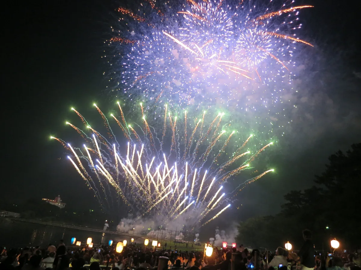 Large fireworks lighting the night sky above a riverbank lined with paper lanterns and spectators