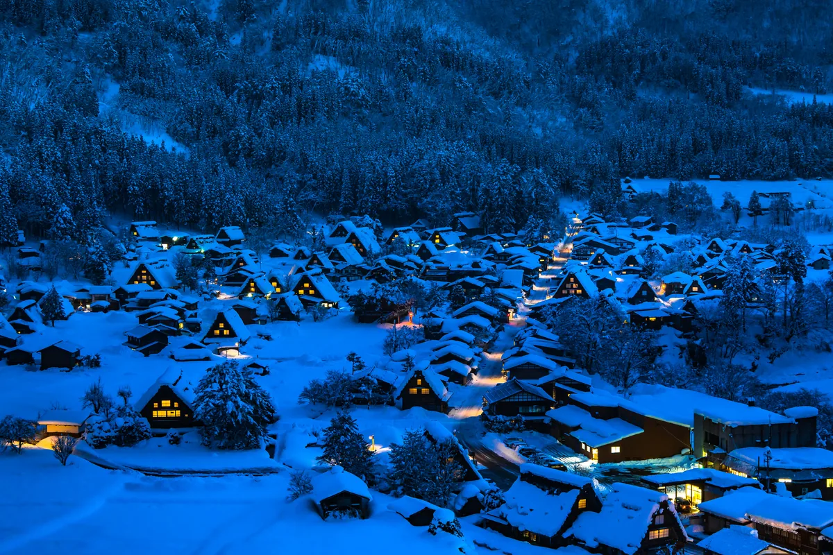 Snow-covered thatched gassho-zukuri houses in a valley at dusk, their windows glowing against deep blue snow