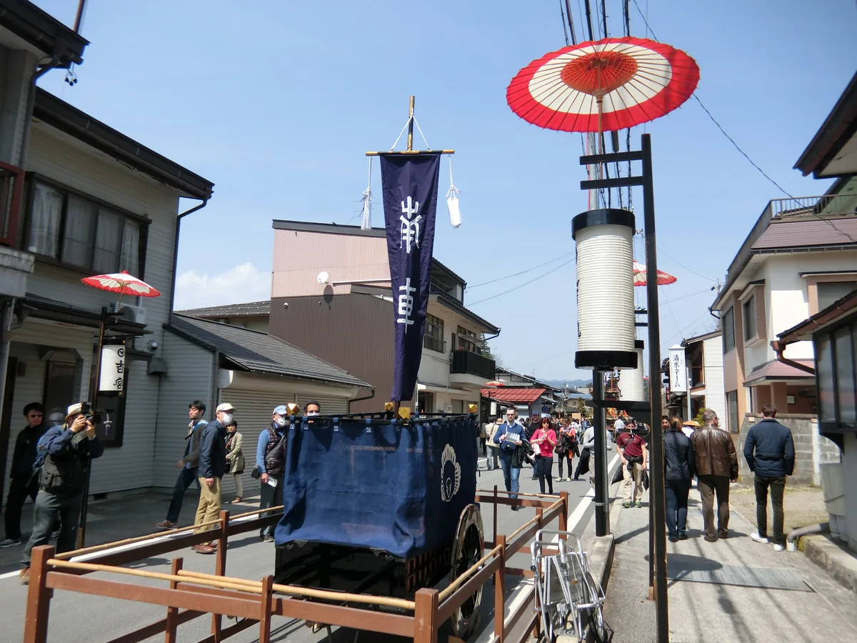 A festival float base draped with a dark banner in a Takayama street, with red paper umbrellas and a tall lantern