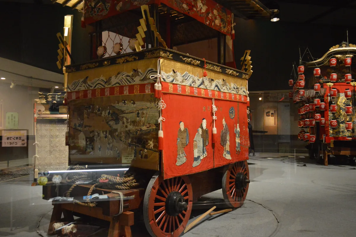 An ornate danjiri festival float with embroidered curtains and drums, displayed in an exhibition hall