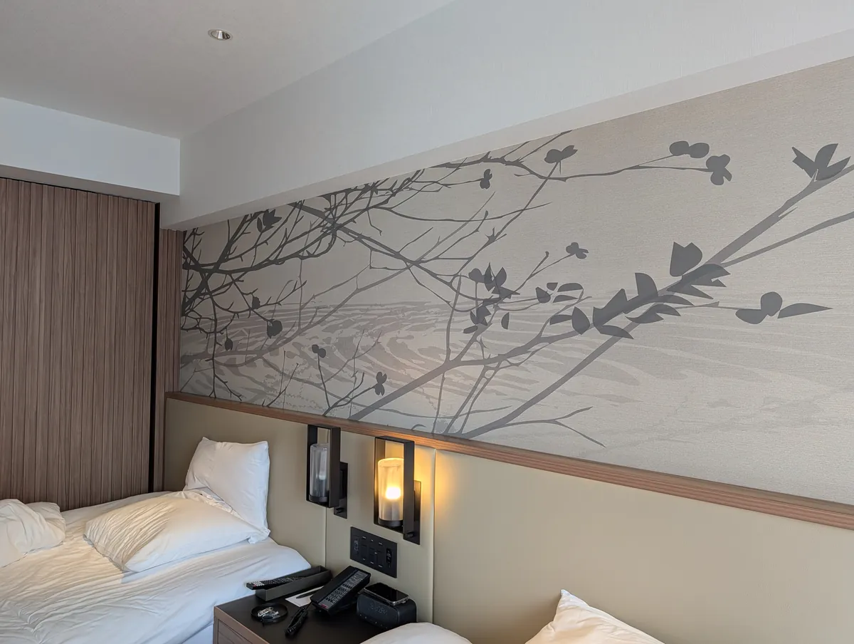 A twin guest room at Courtyard by Marriott Nagoya with two beds side by side, a shared nightstand between them and a large branch-and-leaf artwork covering the headboard wall