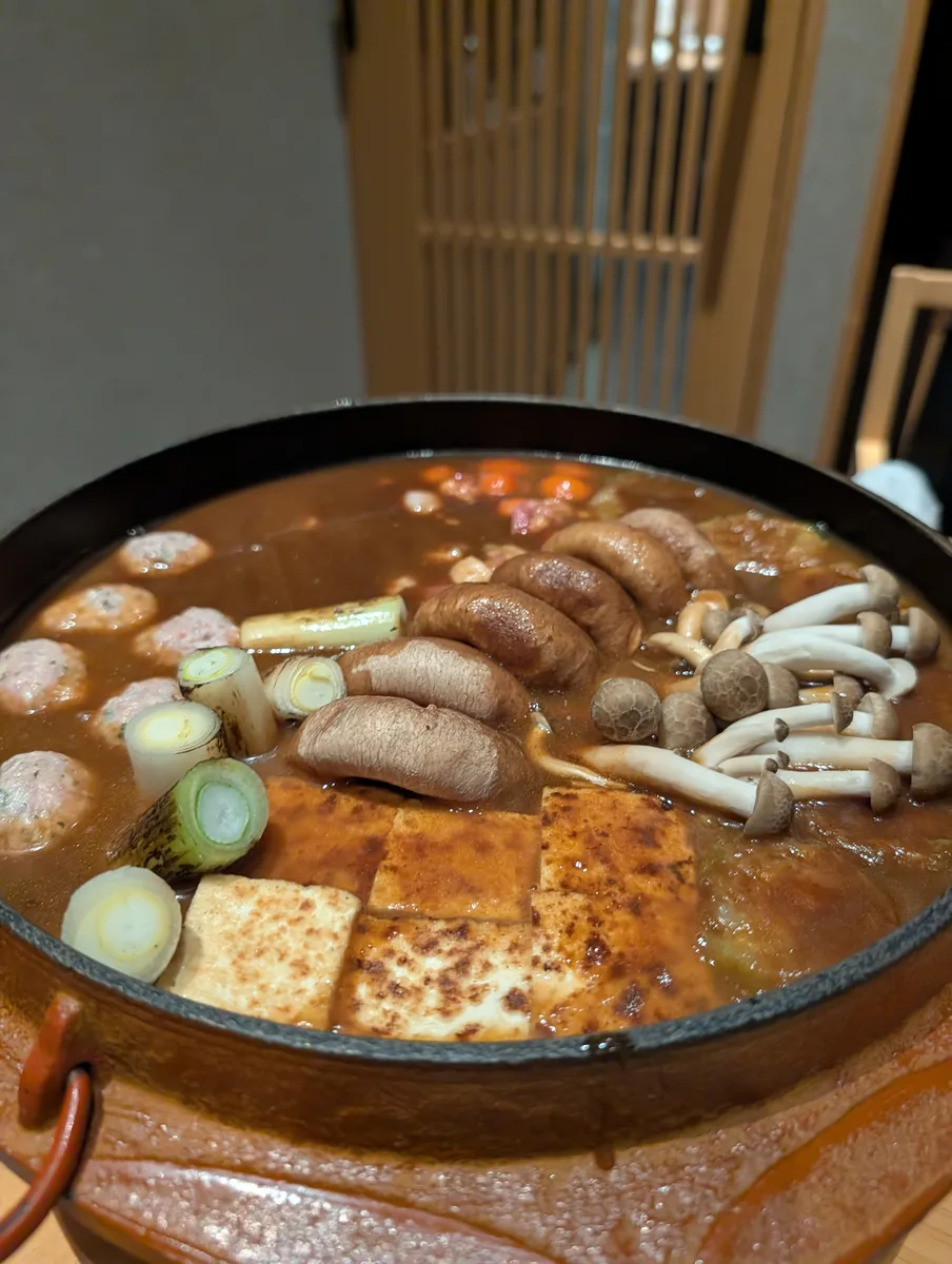 A large clay hotpot of miso-coloured broth filled with meatballs, shiitake and shimeji mushrooms, grilled tofu and sliced leek.