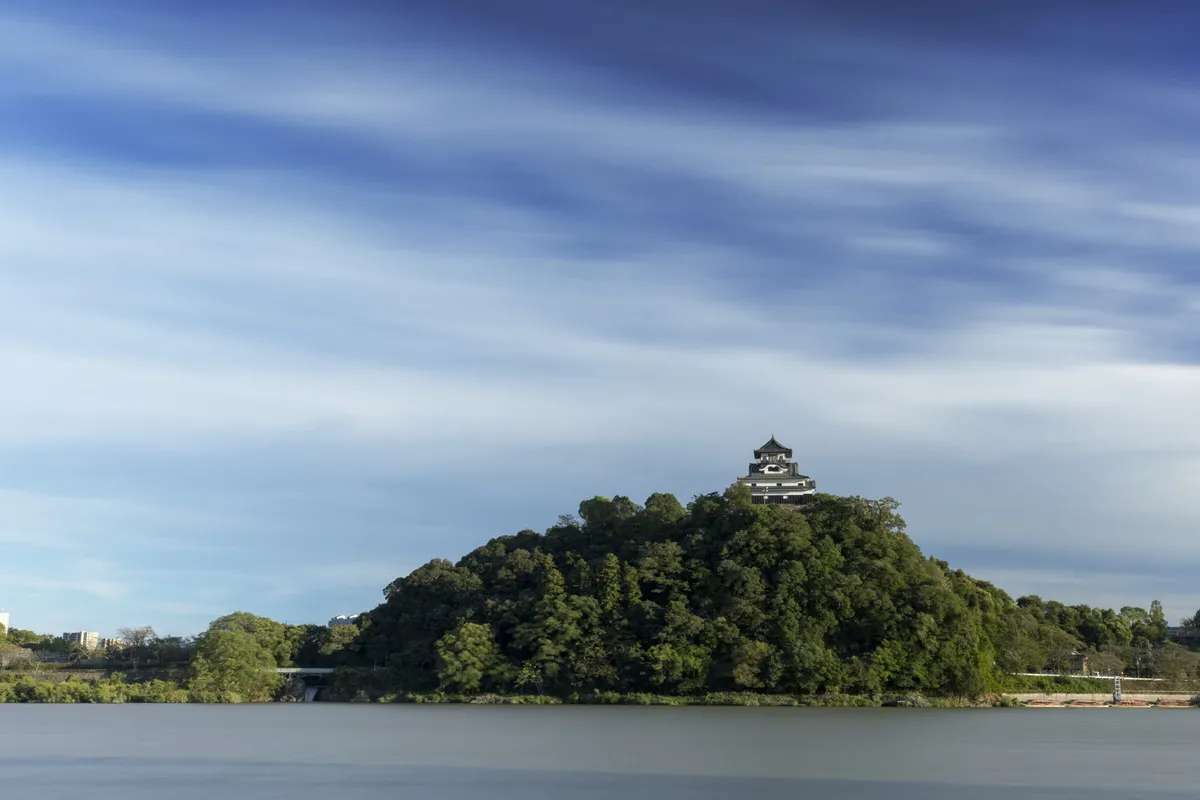 Inuyama Castle keep standing alone on a densely wooded hill above the wide, glassy surface of the Kiso River, under a sky streaked with high cirrus cloud