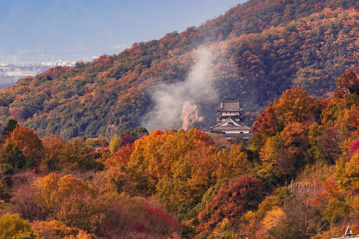 The keep of Inuyama Castle seen at a distance across a hillside covered in red, orange and gold autumn foliage, with pale mist rising from a rocky outcrop on the slope