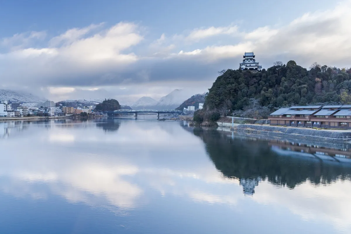 Inuyama Castle above the Kiso River on a still winter morning, with snow lying on the hills behind the town, a steel bridge in the middle distance and cloud reflected in the flat water