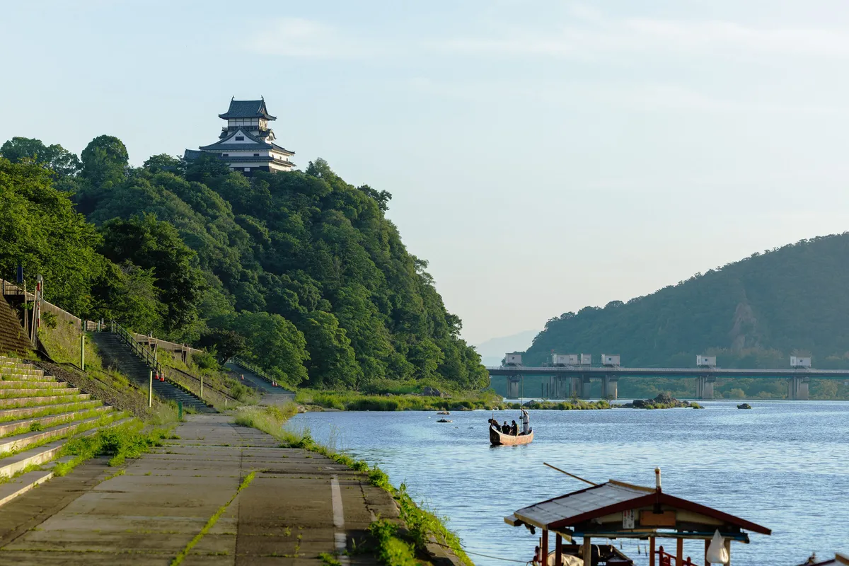 Inuyama Castle on its forested bluff above the Kiso River in summer, with a long wooden boat carrying several standing figures out in mid-river and the roof of a moored covered boat in the foreground