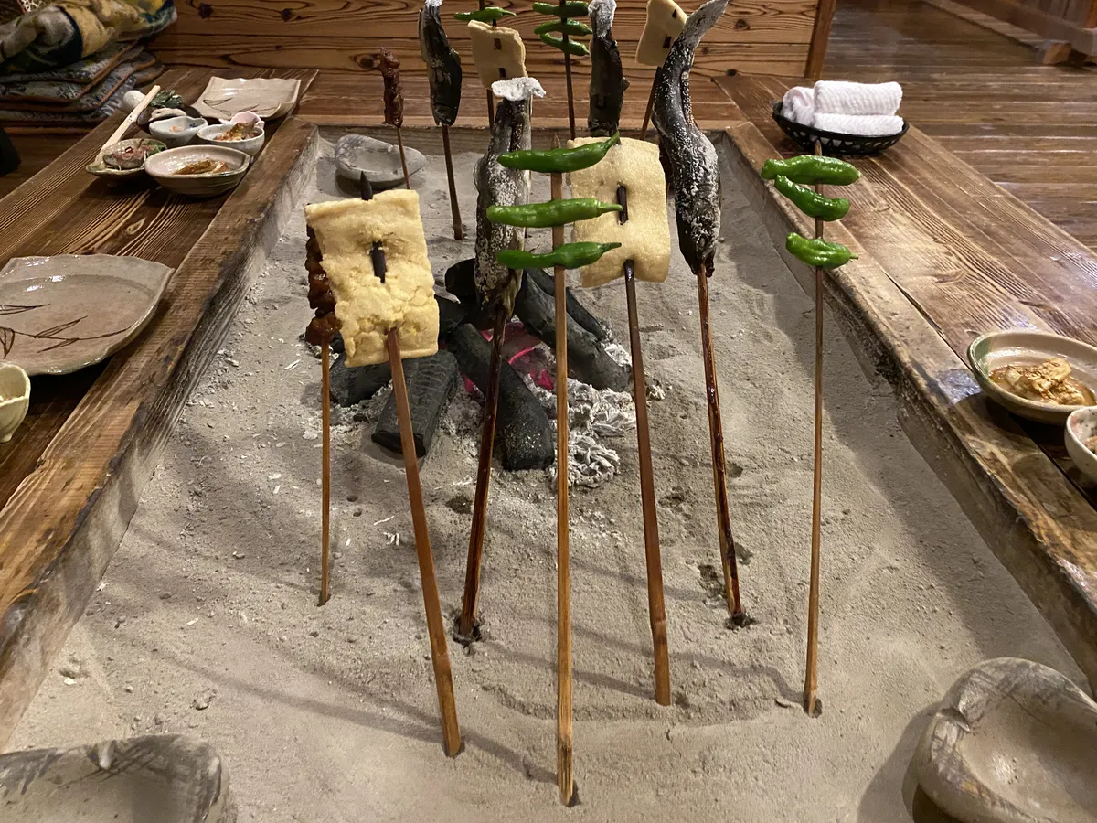 Skewers of river fish, miso-topped tofu and green peppers standing upright in the ash of a sunken charcoal hearth, ringed by a wooden table.
