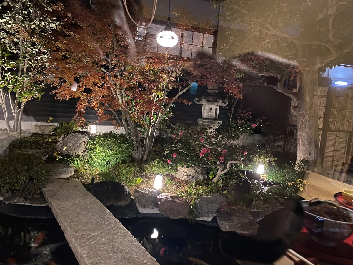 A lit courtyard garden at night seen through glass: a carp pond, a flat stone bridge, moss, a stone lantern and a maple turning red.