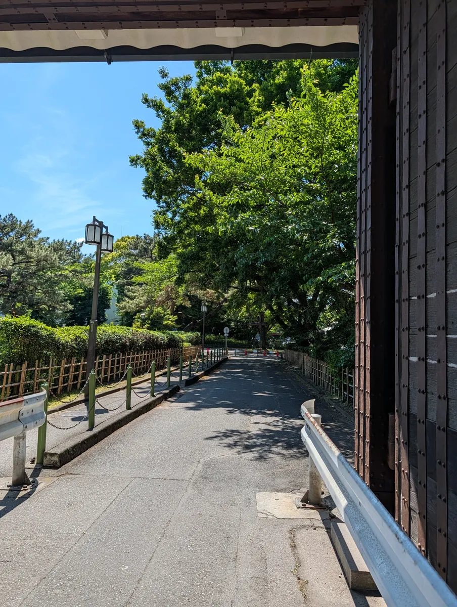 View out from under a Nagoya Castle gate, with the heavy iron-studded timber door filling the right of the frame and a tree-lined road with bamboo fencing and a Japanese-style street lamp beyond
