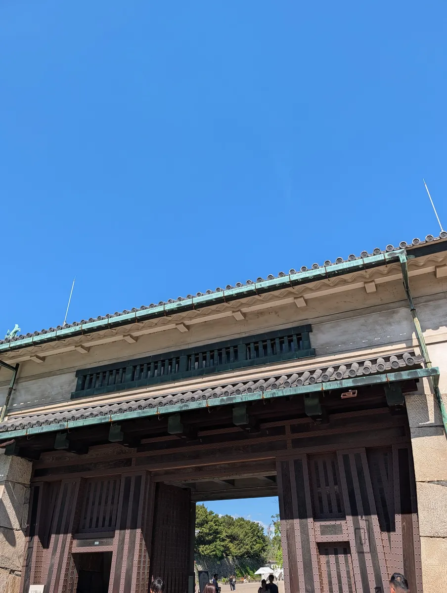 The tall wooden main gate at the entrance to Nagoya Castle, seen from below against a clear blue sky, with a few visitors walking through.