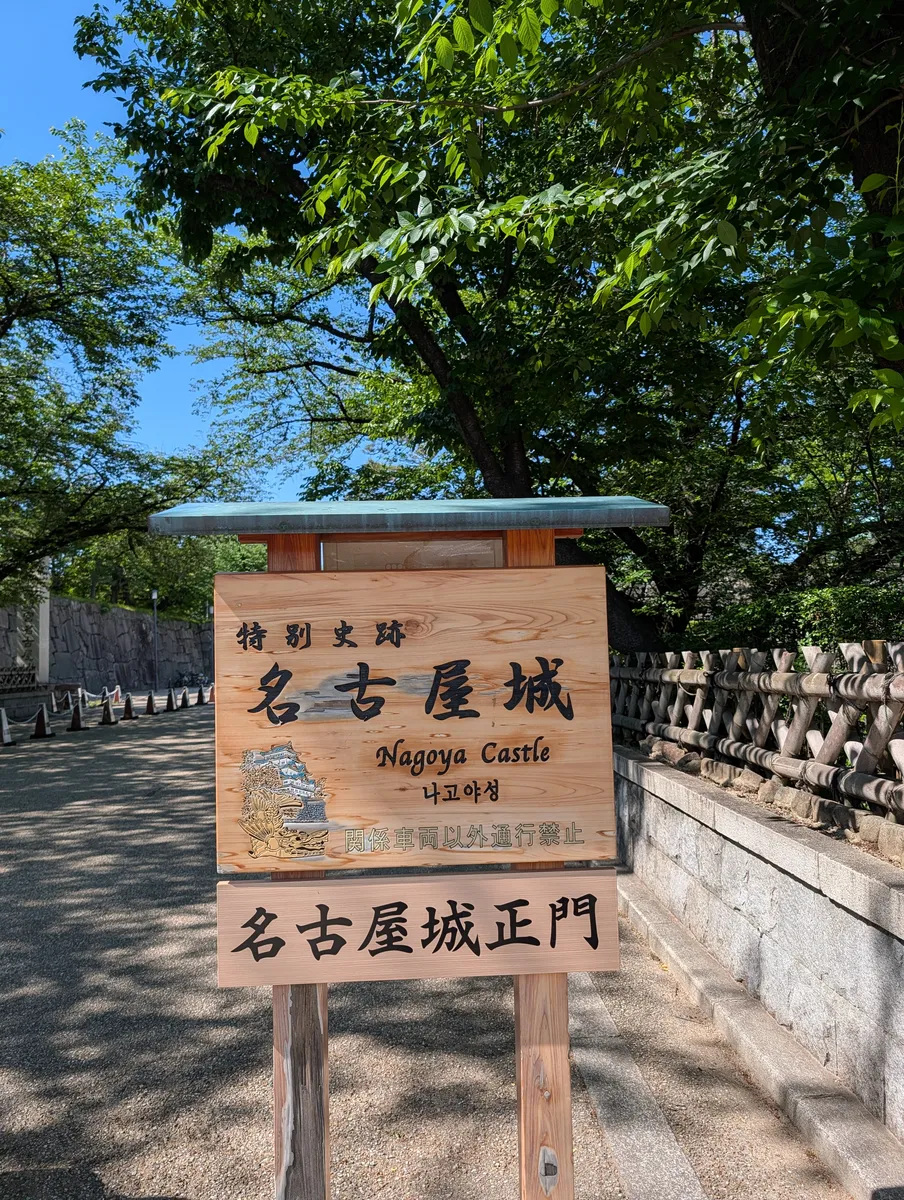 Wooden signboard at a Nagoya Castle entrance carved with the castle name in Japanese, English and Korean above a relief of the keep, with a second board below and a stone wall and bamboo fence behind