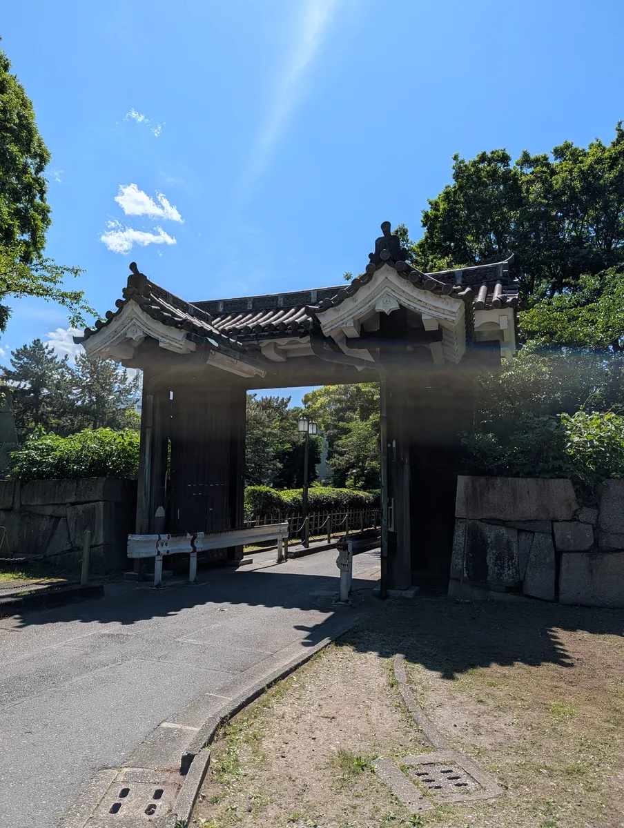A surviving gate at Nagoya Castle seen head-on: a tiled roof over white plaster walls and heavy iron-plated wooden doors, framed by blue sky.