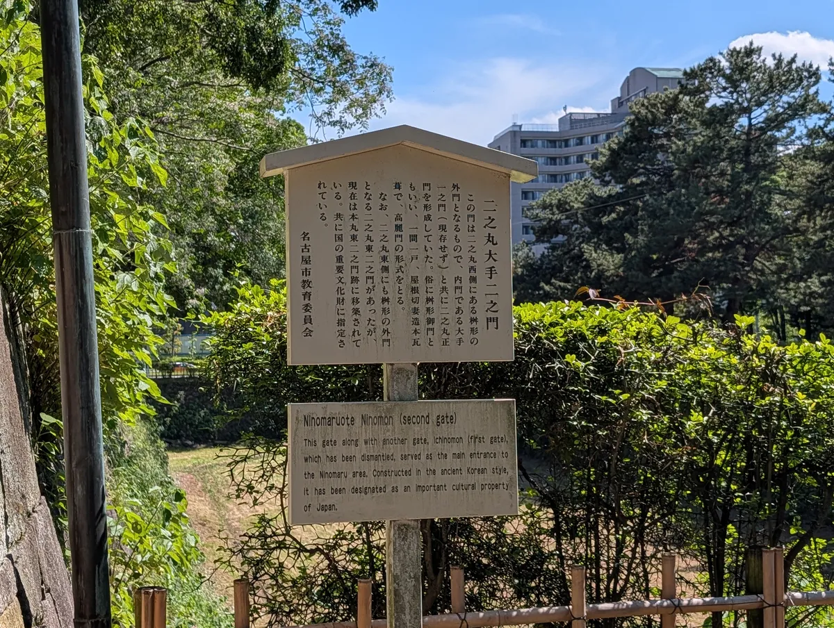 Bilingual interpretive panel at Nagoya Castle headed Ninomaruote Ninomon (second gate), with a Japanese text board above and an English explanation board below, standing among clipped hedges