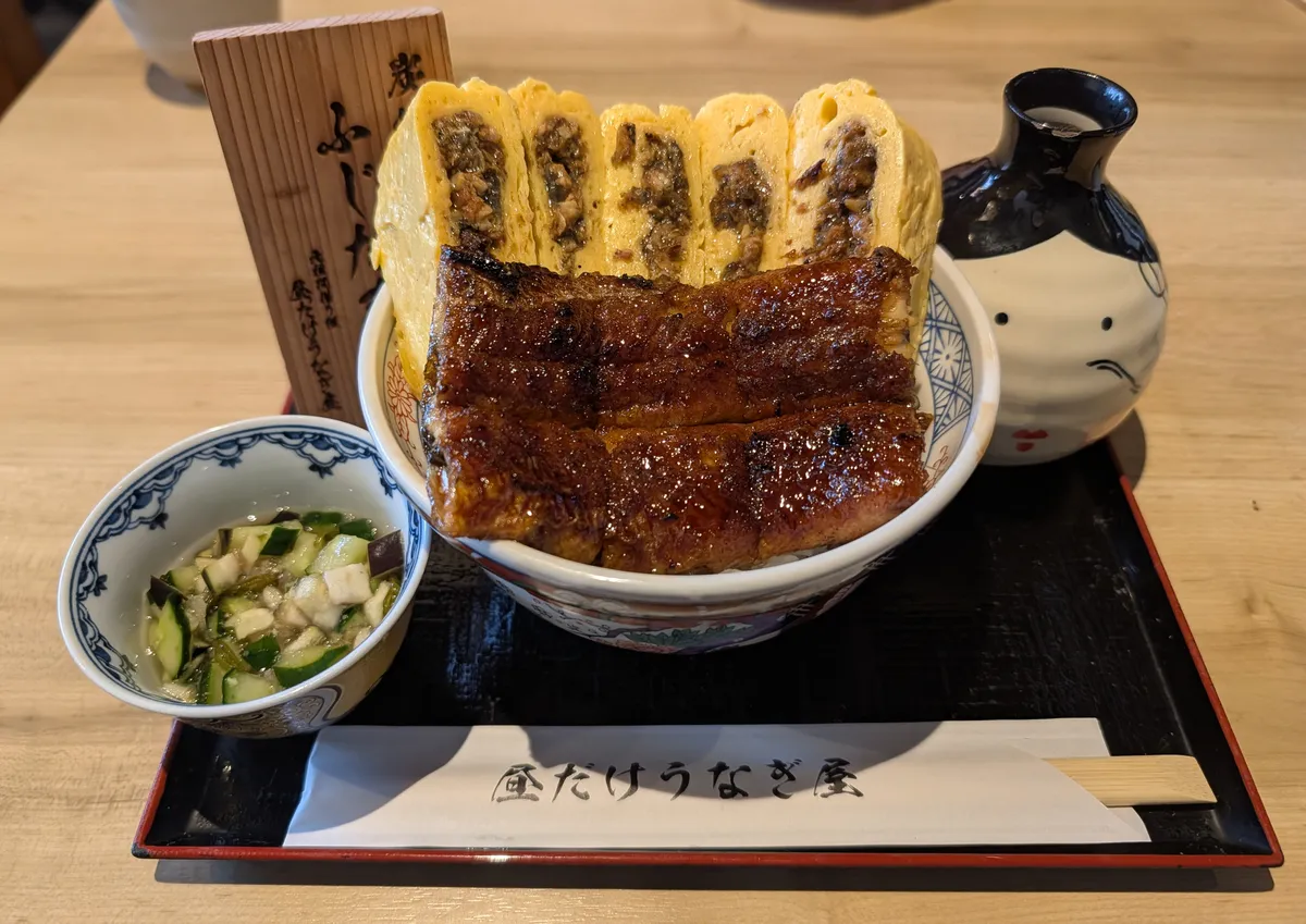 A rice bowl topped with a whole glazed grilled eel fillet and five cut slices of dashimaki omelette rolled around chopped eel, with a wooden menu tag and a pickle dish alongside