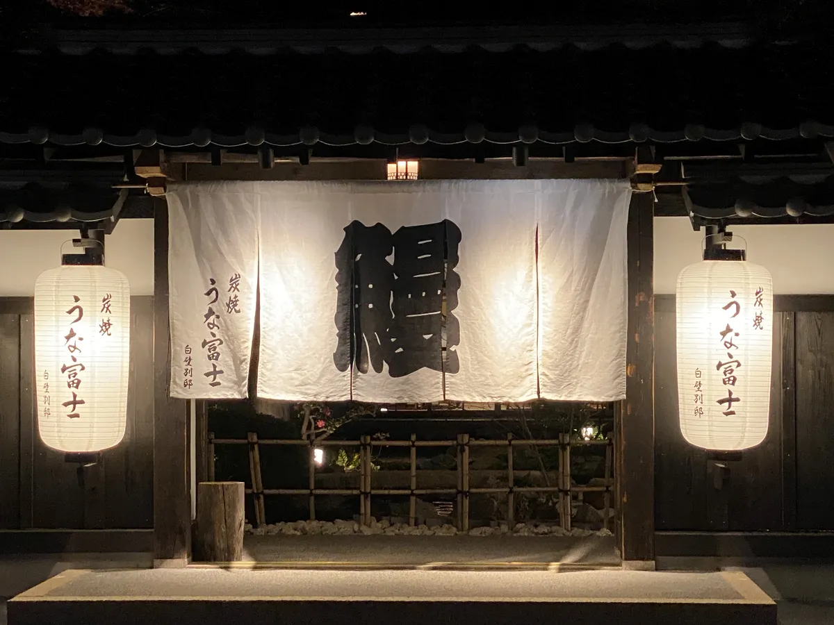 A white noren curtain carrying a single large kanji for eel hangs across a wooden shopfront at night, lit by a paper lantern on each side above a bamboo fence and garden