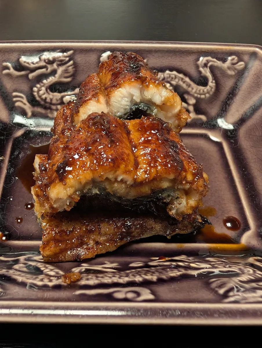 Close-up of three thick pieces of grilled unagi eel stacked on a purple dragon-embossed square plate, the skin lacquered dark with tare sauce