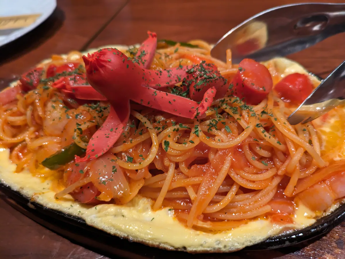 Ketchup-tossed spaghetti with sliced sausage, onion and green pepper on a sizzling iron plate, set over a thin layer of egg.