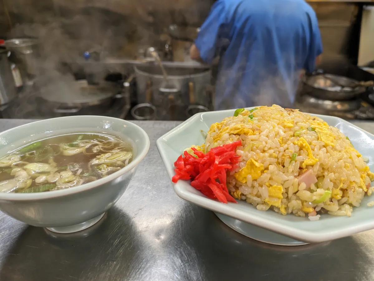 A plate of fried rice with egg, ham and red pickled ginger beside a bowl of clear soup on a steel counter, with a cook working at the steaming range behind