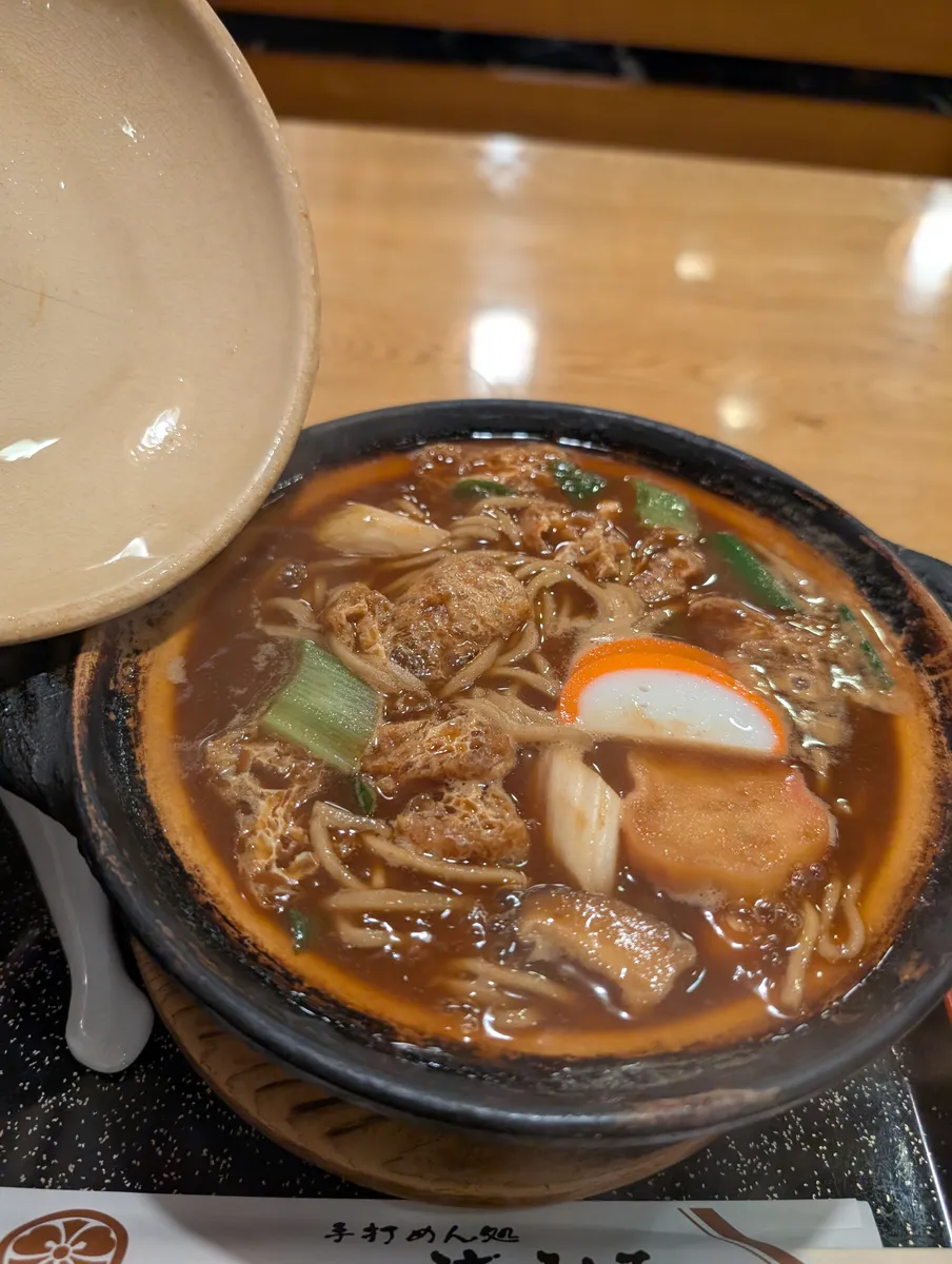 Miso nikomi udon in a black clay pot: thick udon in a dark red-miso broth with fried tofu, fish cake, shiitake and spring onion.