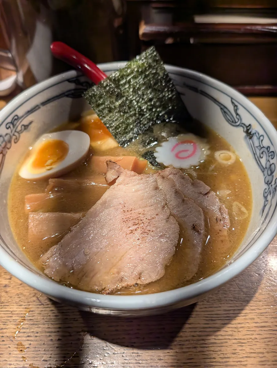 A bowl of ramen in a blue-and-white karakusa bowl: cloudy tan broth topped with two thick chashu slices, a halved marinated egg, menma, a pink-swirled naruto fish cake and a sheet of nori