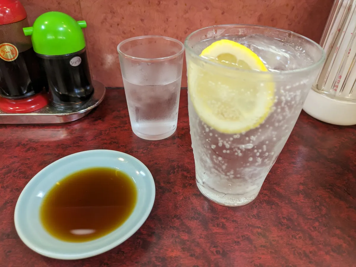 A red lacquered restaurant table set with squeeze bottles labelled chilli oil and soy sauce, a chopstick canister, a glass of iced water, a lemon highball and a small dish of dipping sauce