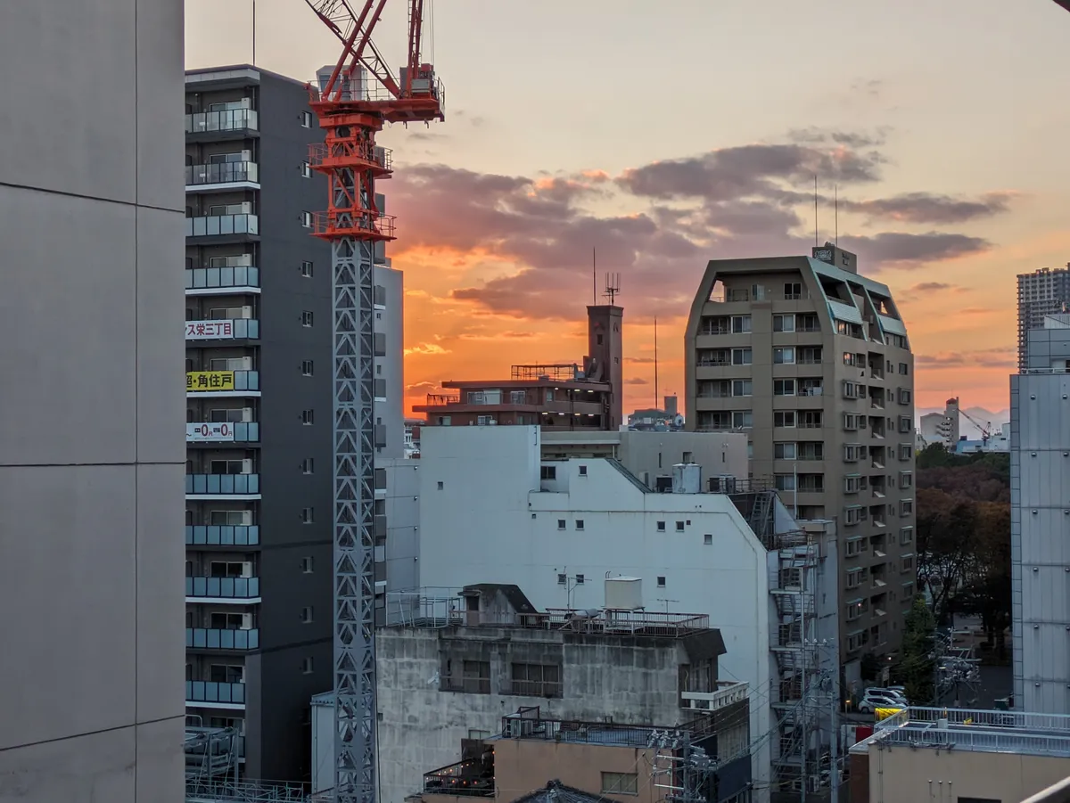 Orange sunset over the packed mid-rise rooftops of central Nagoya, with apartment blocks, a construction crane and a strip of park trees on the right