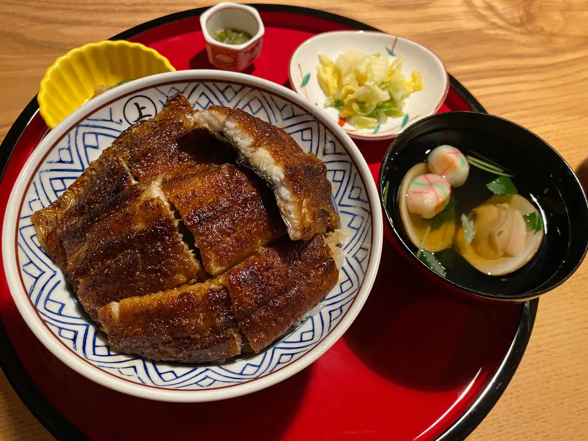 An eel rice bowl in blue-and-white porcelain on a red lacquer tray, with pickled cabbage, a clear soup holding clams and pink temari-fu, and a small pot of sansho pepper