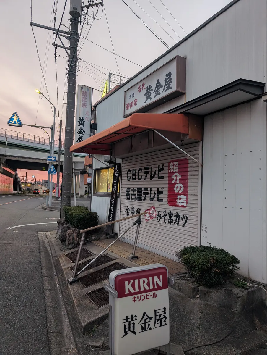 A quiet Nagoya back street at dawn in late December, with a shuttered shopfront, power lines and an elevated expressway under a pink sky.