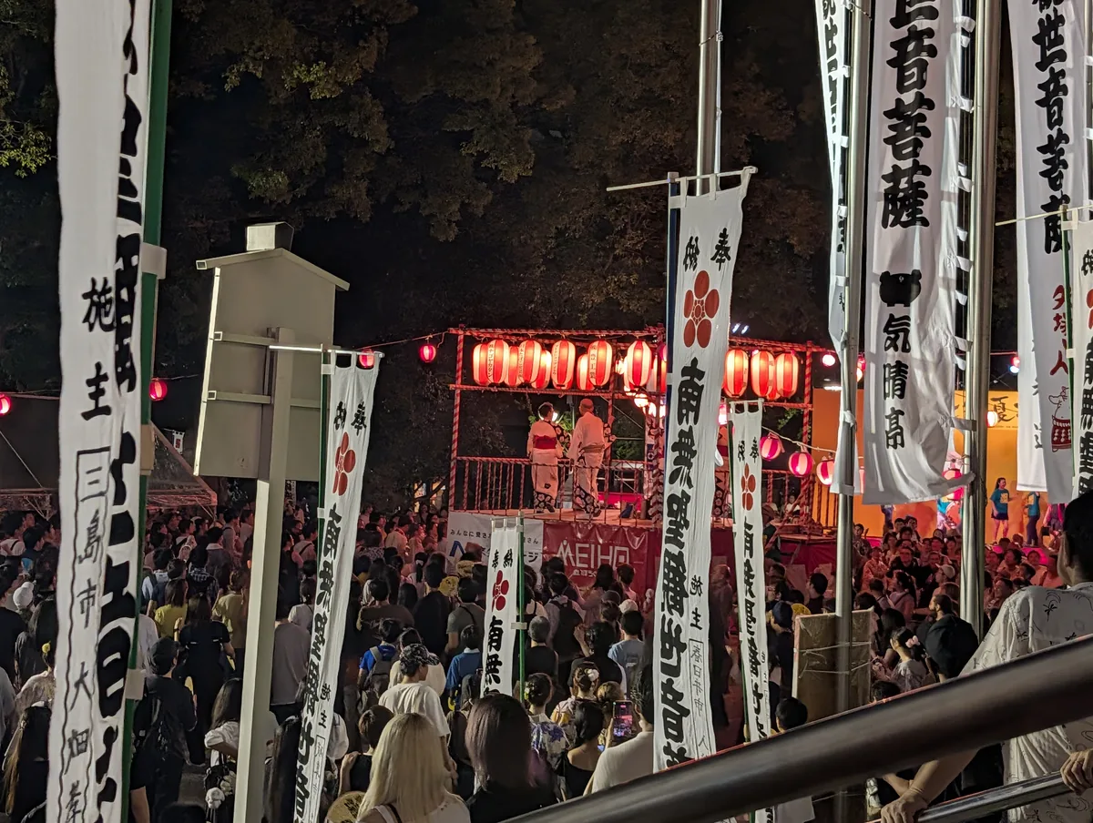 Red lanterns strung around a wooden yagura tower at a bon odori, with a crowd and tall white banners around it at night