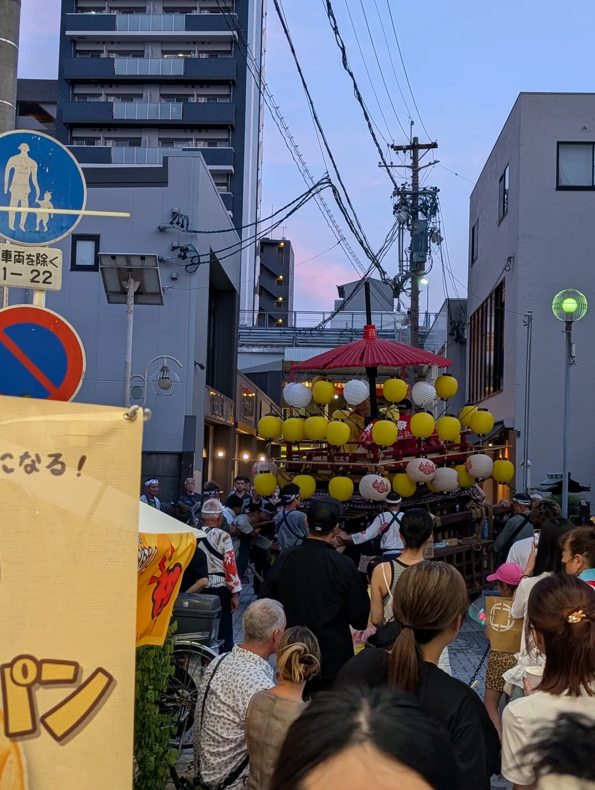 A small festival float decked with yellow and white paper lanterns and a red parasol on a narrow street in Osu