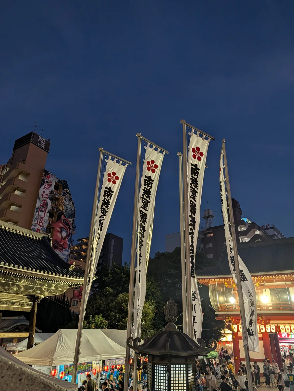 Tall white banners against a deep blue evening sky above a row of lit food stalls on temple grounds in Nagoya