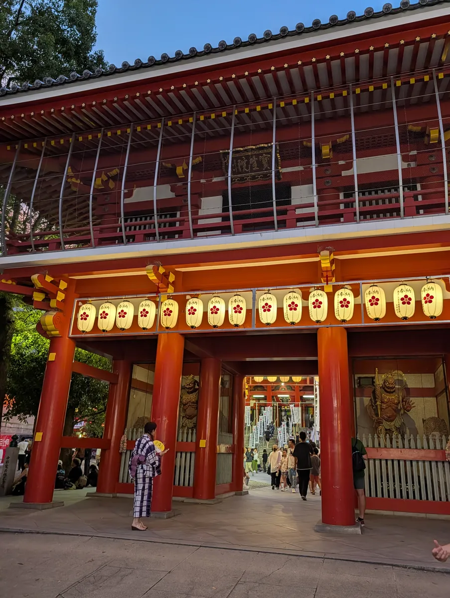 A visitor in a navy-and-white checked yukata, seen from behind holding a round paper fan, standing in front of the lantern-hung vermilion gate at Osu Kannon