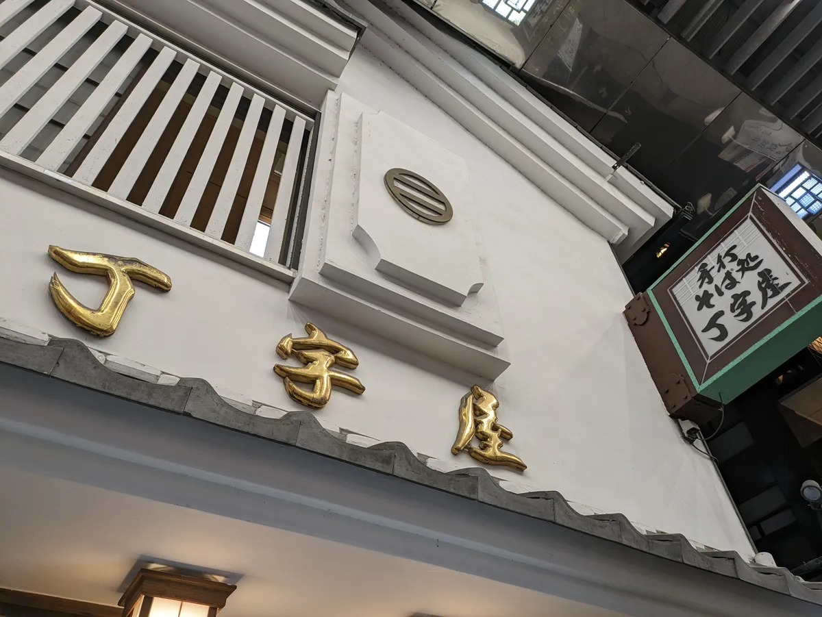 Gilded kanji characters mounted across the white plaster gable of a traditional shopfront in Osu, with a grey tiled ridge below, a slatted upper window and a lantern-style hanging sign to the right